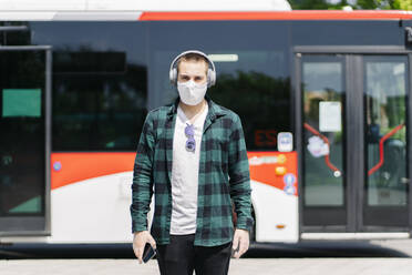 Porträt eines jungen Mannes mit Schutzmaske, der mit Kopfhörern vor einem Bus Musik hört, Spanien - DGOF01021