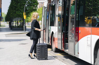Geschäftsfrau mit Schutzmaske vor einem Bus mit Trolley-Tasche, Spanien - DGOF01027