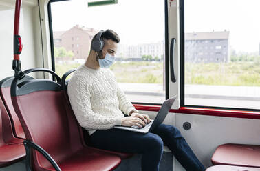 Junger Mann mit Schutzmaske sitzt in einem öffentlichen Bus und benutzt Kopfhörer und Laptop, Spanien - DGOF01043