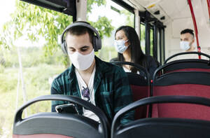 Portrait of young man with headphones wearing protective mask in public bus using cell phone, Spain - DGOF01047