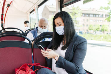 Businesswoman wearing protective mask in public bus looking at cell phone, Spain - DGOF01068