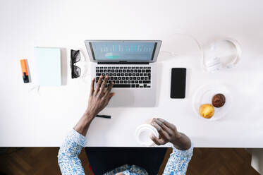 Man holding coffee while using laptop at desk in home office - JCMF00765