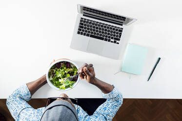Young man eating salad while sitting with laptop at home office desk - JCMF00776