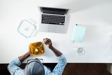 Junger Mann isst Suppe aus einem Behälter, während er mit einem Laptop am Schreibtisch im Büro sitzt - JCMF00777