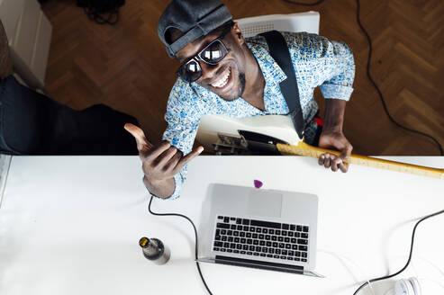 Portrait of smiling young man showing shaka sign while sitting with guitar and laptop at desk - JCMF00781