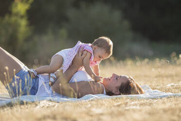 Glückliche Frau, die ein kleines Mädchen trägt, während sie auf einer Decke auf einer Wiese in der Sonne liegt - SNF00229