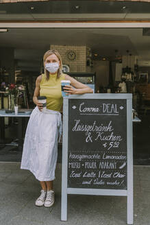 Frau mit Gesichtsmaske und Coffee to go in der Hand, während sie sich an ein Schild mit Corona-Sonderangeboten lehnt - MFF05679