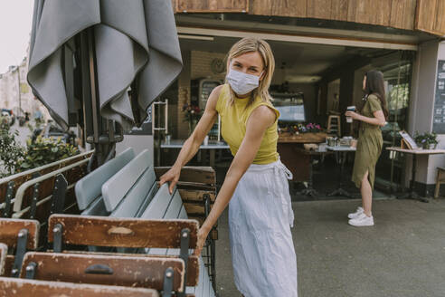 Cafe owner wearing face mask putting up tables and chairs - MFF05680