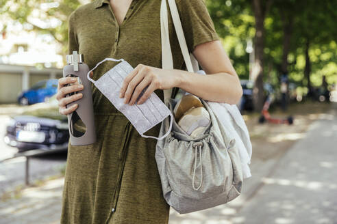 Close-up of woman holding a face mask and drinking bottle in her hand while walking to her health club - MFF05685