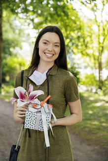 Porträt einer glücklichen Frau in der Natur, die eine Lilienblüte und ein Geschenk hält - MFF05698