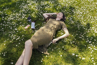Glückliche Frau, die ihre Freizeit genießt, während sie mit Gänseblümchen neben der Gesichtsmaske im Gras liegt - MFF05706