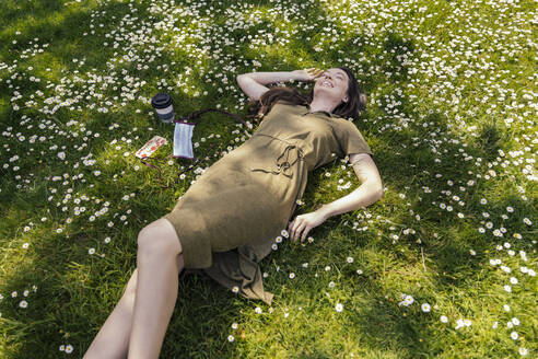 Happy woman enjoying her free time while lying on grass with daisies next to face mask - MFF05706