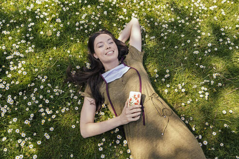 Frau mit heruntergelassener Gesichtsmaske genießt ihre Freizeit, während sie im Gras mit Gänseblümchen liegt - MFF05708