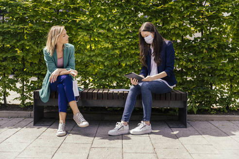 Two businesswomen sitting on bench outside and keeping their distance while wearing face mask - MFF05724