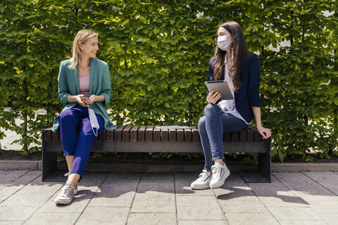Two businesswomen sitting on bench outside and keeping their distance while wearing face mask stock photo
