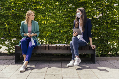 Two businesswomen sitting on bench outside and keeping their distance while wearing face mask - MFF05725