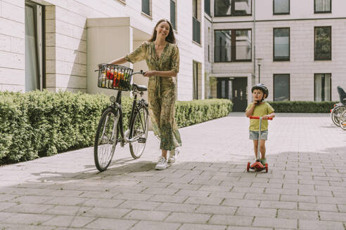 Lächelnde Mutter mit Fahrrad und Sohn mit Motorroller in der Stadt - MFF05727