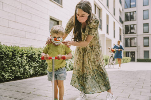 Woman putting on face mask on son with scooter in the city - MFF05729