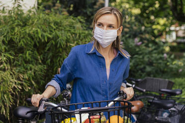 Woman wearing face mask with bicycle and shopped groceries in urban area - MFF05736