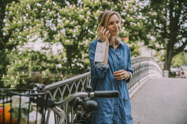 Frau auf einer Brücke mit Gesichtsmaske und Fahrrad beim Telefonieren - MFF05755