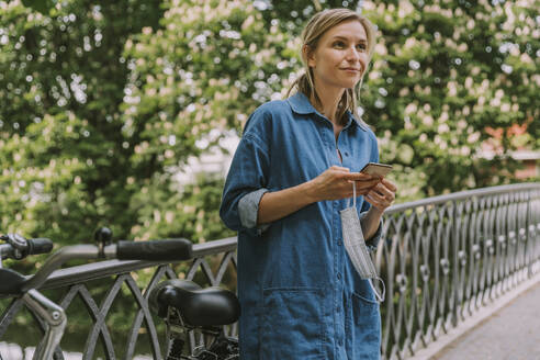 Frau auf einer Brücke mit Gesichtsmaske, Smartphone und Fahrrad - MFF05756