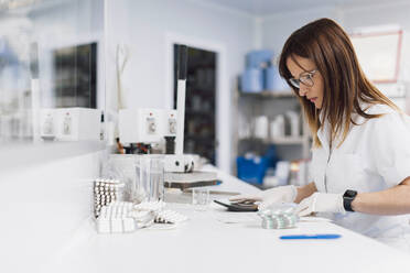 Confident mature female healthcare worker with pills and calculator at desk in laboratory - OCAF00491