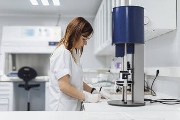 Confident mature female healthcare worker writing data at laboratory desk - OCAF00515