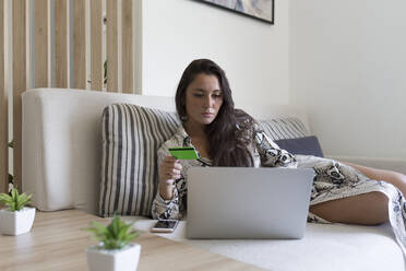 Schöne junge Frau hält Kreditkarte für Online-Shopping durch Laptop, während auf dem Sofa zu Hause liegen - JPTF00491