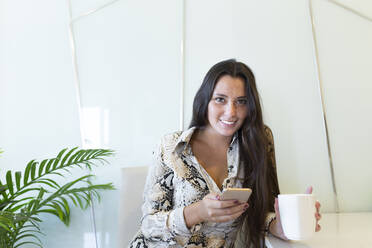 Portrait of smiling young woman holding coffee cup and mobile phone while sitting at home - JPTF00492