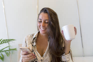 Lächelnde schöne junge Frau mit Sommersprossen, die ein Smartphone benutzt und eine Kaffeetasse zu Hause hält - JPTF00494