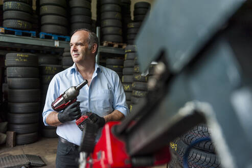 Ein älterer Unternehmer hält einen Schlagschrauber in der Hand, während er in einem Reifengeschäft wegschaut - DIGF11564