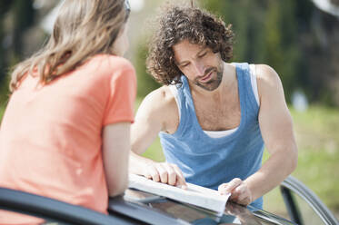 Confident handsome man reading map on car roof with woman during summer road trip - DIGF11613