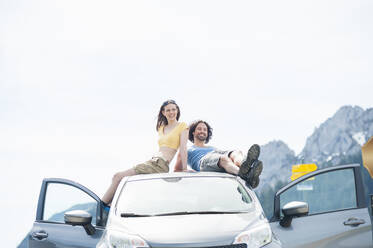 Mid adult couple relaxing on car roof against clear sky during summer road trip - DIGF11616