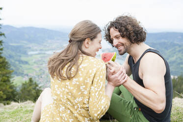 Glückliches erwachsenes Paar, das auf einem Berggipfel sitzend ein Stück Wassermelone genießt - DIGF11622