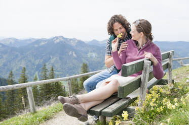 Frau sieht Mann an, der an einer Blume riecht, während er auf einer Bank in den Bergen sitzt, gegen den klaren Himmel - DIGF11630