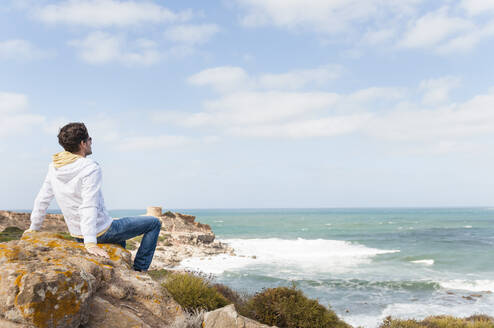 Mann entspannt sich auf einem Felsen an der Küste und schaut aufs Meer, Sardinien, Italien - DIGF11695