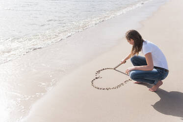 Frau hockt am Strand und kratzt mit einem Holzstab ein Herz in den nassen Sand, Sardinien, Italien - DIGF11701