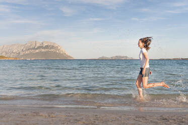 Happy woman running at seashore splashing with water, Sardinia, Italy - DIGF11711