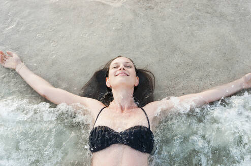 Porträt einer glücklichen Frau im Bikini, die am Meeresufer liegt und die Brandung genießt, Sardinien, Italien - DIGF11716