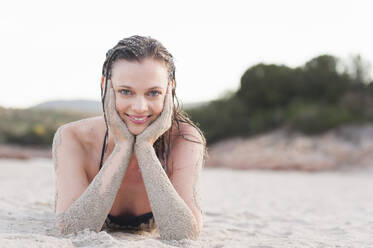 Porträt einer lächelnden Frau, die mit dem Kopf in den Händen am Strand liegt, Sardinien, Italien - DIGF11719