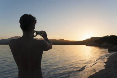 rückansicht eines Mannes, der mit seinem Handy den Sonnenuntergang fotografiert, Sardinien, Italien - DIGF11733