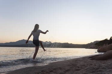 Rückenansicht einer Frau, die bei Sonnenuntergang am Meeresufer im Wasser plantscht, Sardinien, Italien - DIGF11735