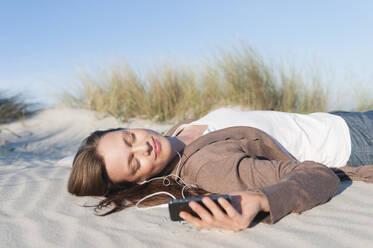 Portrait of woman lying on beach dune listening music with earphones and smartphone, Sardinia, Italy - DIGF11759