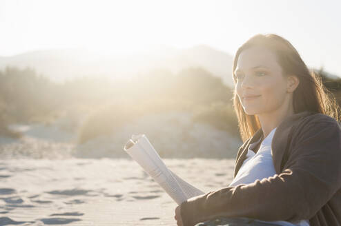 Portrait of smiling woman with magazine lying on the beach looking at distance, Sardinia, Italy - DIGF11760