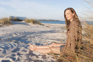 Portrait of woman sitting at beach dunes listening music with earphones, Sardinia, Italy - DIGF11765