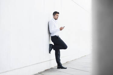 Businessman using smartphone leaning on white wall - DIGF11776