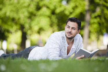Young man reading book lying on blanket in park - DIGF11787