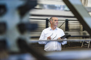 Älterer Geschäftsmann mit Tablet in einer Fabrikhalle, der sich umschaut - DIGF11826