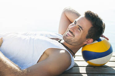 Porträt eines lächelnden jungen Mannes mit Ball, der auf einem Steg vor einem See liegt - DIGF11934