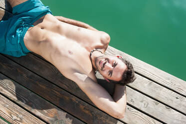 Porträt eines lächelnden jungen Mannes in Badeshorts beim Sonnenbaden auf einem Steg - DIGF11944